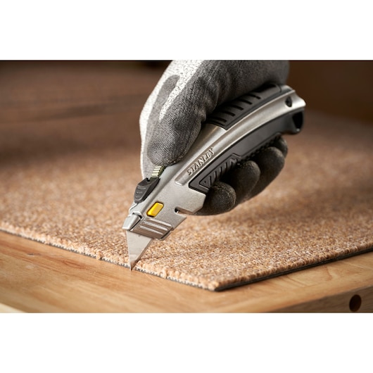 Photo du couteau utilitaire STANLEY 0-10-788 coupant un tapis, tenu par une main gantée.
