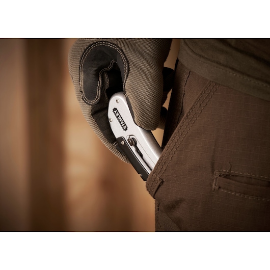 Couteau utilitaire pliant STANLEY 0-10-810 tenu par une main gantée, placé dans une poche de pantalon marron, gros plan.