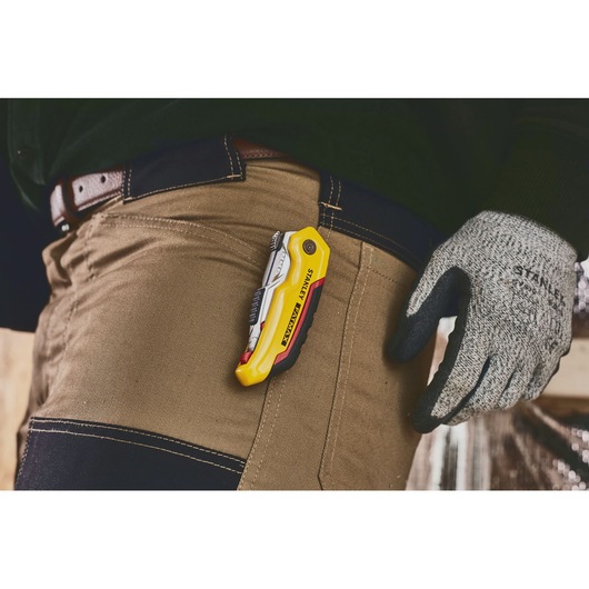 Couteau utilitaire STANLEY 0-10-825 accroché à un pantalon de travail marron, personne portant des gants.