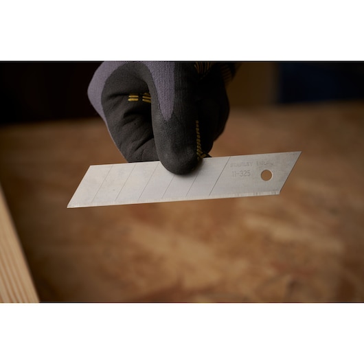 Photographie de la lame de cutter STANLEY 0-11-325 tenue par une main gantée, lignes segmentaires visibles.