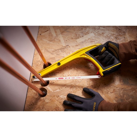 Photo de la scie à métaux STANLEY 0-20-108 à cadre jaune découpant un tuyau en cuivre, tenue avec des gants de travail.