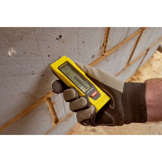 STANLEY 0-77-030 télémètre laser tenu par une main gantée contre un mur de briques, produit en usage.