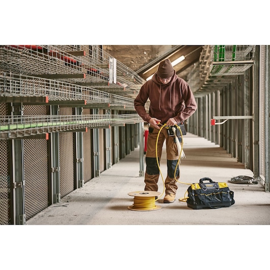STANLEY 0-84-004 gereedschapstas geel en zwart op de grond naast medewerker met gele kabelhaspel in industriële omgeving.
