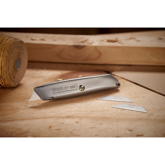 Un couteau utilitaire en métal Stanley 99E posé sur une surface en bois, avec deux lames de rechange à proximité. L'arrière-plan montre des matériaux et outils de menuiserie.