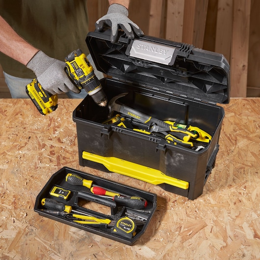 STANLEY 1-70-316 zwarte en gele gereedschapskist met diverse handgereedschappen en open deksel, persoon houdt boormachine vast.