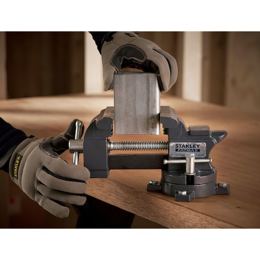 Foto van STANLEY 1-83-065 bankschroef die een metalen stuk op een werkbank klemt, monteur met handschoenen.