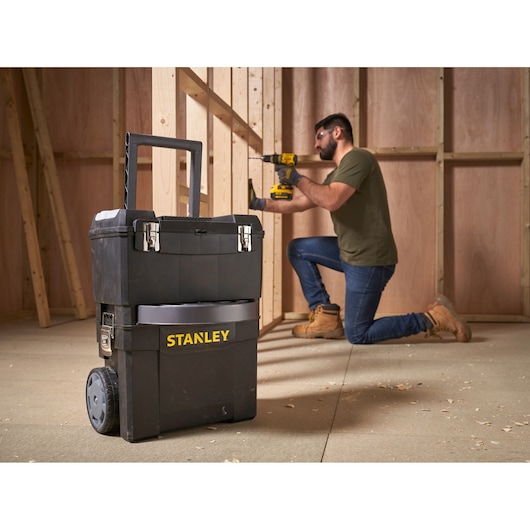 STANLEY 1-93-968 mobiele gereedschapskist met wielen in werkplaats, werker gebruikt elektrisch gereedschap op de achtergrond.