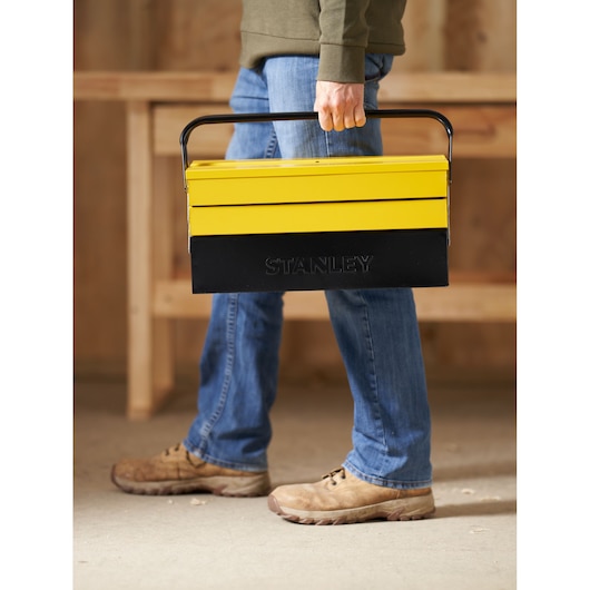 Boîte à outils cantilever en métal jaune et noir Stanley 1-94-738 portée par une personne en intérieur.