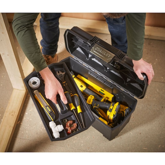 Een persoon opent een zwarte STANLEY gereedschapskist met verschillende handgereedschappen zoals schroevendraaiers, een moersleutel, een stanleymes, tape en koppelingen. De gereedschapskist heeft diverse compartimenten voor organisatie en bevat geel en zwart gereedschap.