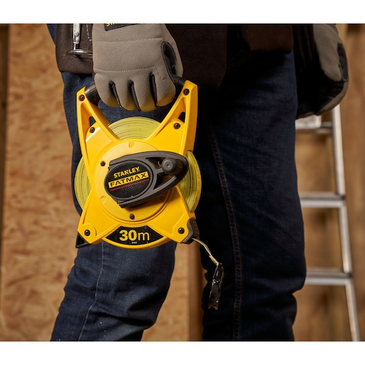 STANLEY 2-34-814 FATMAX 30m open rolmaat vastgehouden door een gehandschoende hand, werkplaats op de achtergrond.