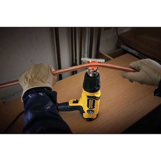 STANLEY FME670K pistolet à air chaud utilisé pour plier un tube en cuivre, personne gantée tenant le tube sur un établi.
