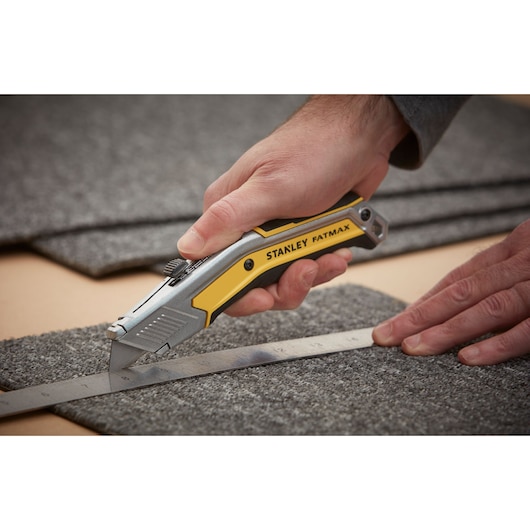 Photo du couteau utilitaire STANLEY FATMAX FMHT0-10288 découpant de la moquette le long d'une règle métallique.