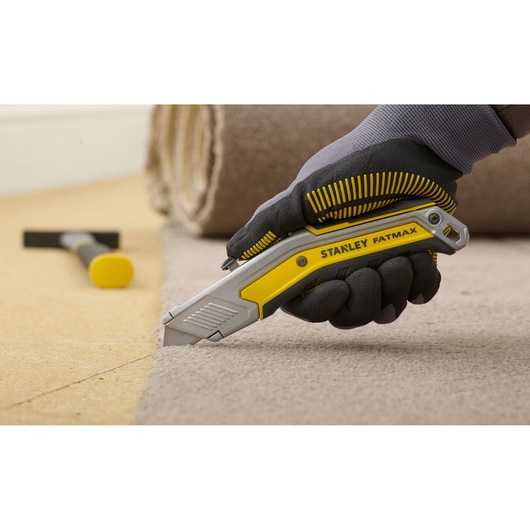 Cutter STANLEY FMHT0-10288 découpant de la moquette, poignée ergonomique jaune et argent, main gantée tenant l’outil.