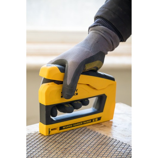 STANLEY FMHT0-80551 tacker in geel en zwart, vastgehouden door een gehandschoende hand, drukt gaas op hout.