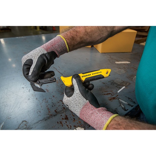 Foto van STANLEY FMHT10358-0 gereedschapsmes met gele handgreep vastgehouden door gehandschoende handen op metalen tafel.