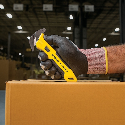STANLEY FMHT10358 hobbymes met gele handgreep, vastgehouden met handschoen, snijdt kartonnen doos.