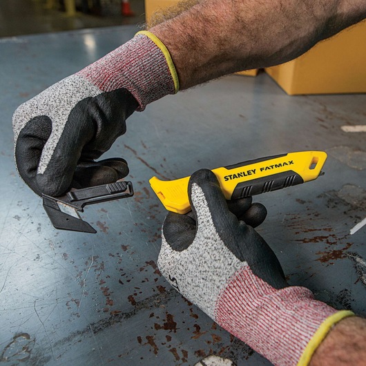 Stanley FMHT10358 gereedschapsmes in handschoenen vastgehouden, gele handgreep, geopend lemmetvak, industriële setting.