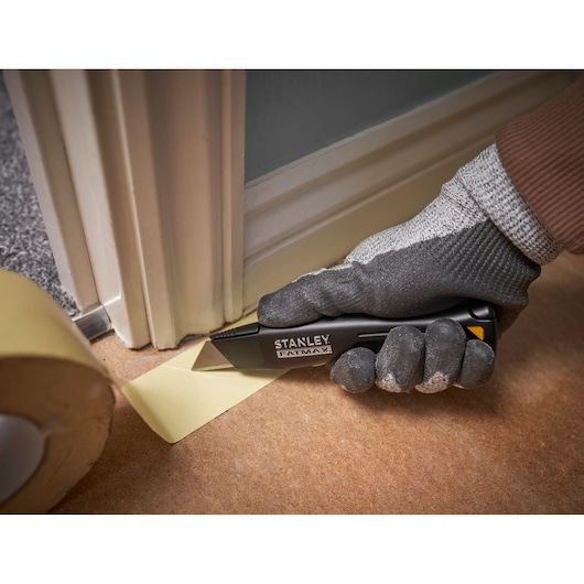 Couteau utilitaire STANLEY FMHT10500-0 coupant du ruban de masquage près d’un cadre de porte, main gantée tenant le couteau.
