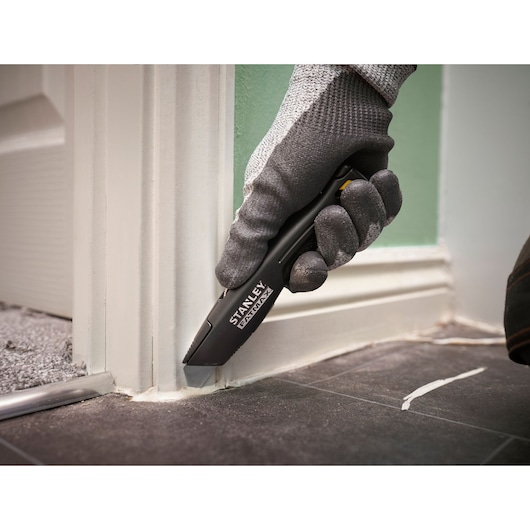 Photo du couteau utilitaire STANLEY FMHT10500-0 coupant du mastic au coin d’un cadre de porte sur carrelage noir.