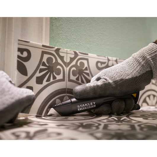STANLEY FMHT10500-0 couteau utilitaire tenu par une main gantée, coupant du joint de carrelage à motif, vue latérale.