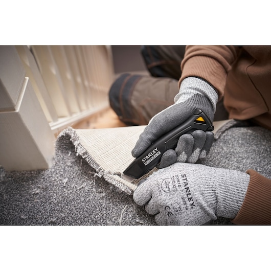 Photo du couteau utilitaire STANLEY FMHT10500-0 coupant un tapis, mains gantées tenant le couteau et le tapis.