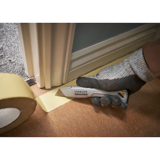STANLEY FMHT10501-0 couteau utilitaire coupant du ruban près d’un cadre de porte, photo en gros plan avec main gantée.