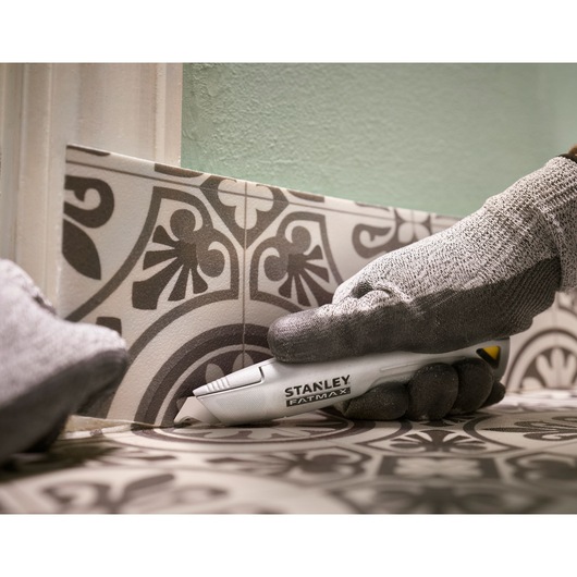 Photographie du couteau utilitaire STANLEY FMHT10501-0 découpant un carrelage avec une main gantée, vue rapprochée.
