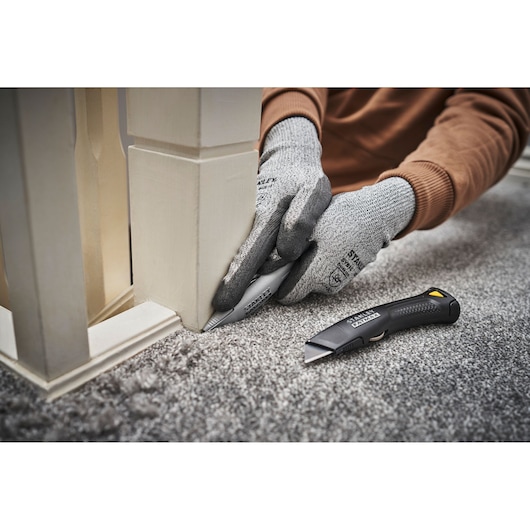 Photo du couteau utilitaire STANLEY FMHT10501-0 utilisé pour couper de la moquette le long d'un poteau blanc, avec gants.
