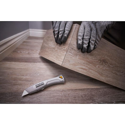 Cutter STANLEY FMHT10501-0 sur parquet, à côté de mains gantées posant un panneau de sol, vue en angle.
