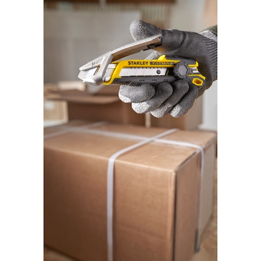 STANLEY FMHT10592-0 couteau utilitaire tenant dans une main gantée, jaune et noir, cartons en arrière-plan.