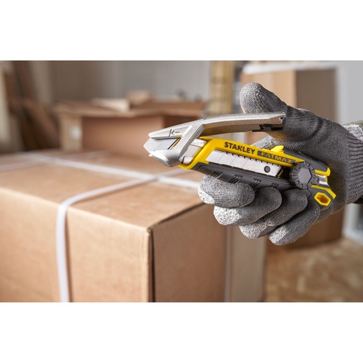 Cutter STANLEY FMHT10592-0 tenu par une main gantée, coupant une sangle de carton dans un entrepôt.