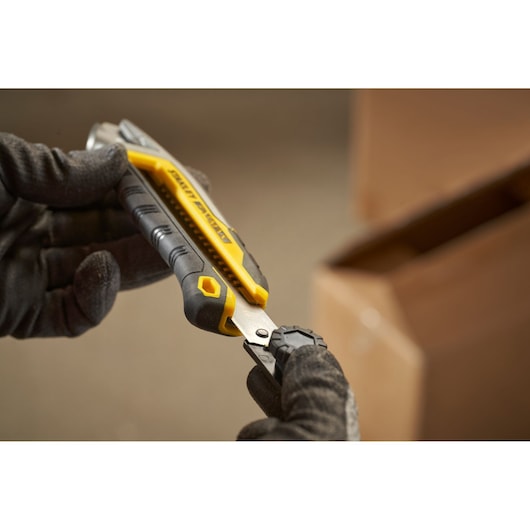 STANLEY FMHT10592-0 couteau utilitaire à poignée jaune et noire tenu par des mains gantées près d'une boîte carton.