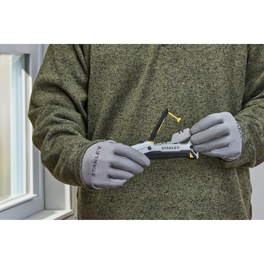 Persoon met grijze Stanley-handschoenen en een groene trui houdt en demonstreert een Stanley FMHT10722-0 afbreekmes voor een raam.