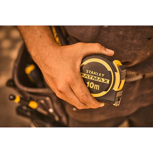 Foto van STANLEY FATMAX 10m rolmaat FMHT33005-0 in hand, met gereedschapsriem en gereedschap op de achtergrond.