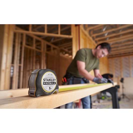 STANLEY FMHT38214-0 FATMAX rolmaat op houten plank, bouwscène met persoon op de achtergrond.