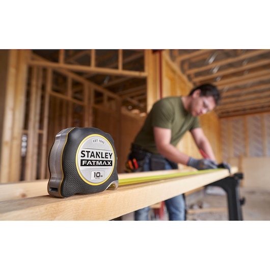 STANLEY FATMAX 10m rolmaat FMHT38232-0 op houten balk, bouwvakker meet op de achtergrond.