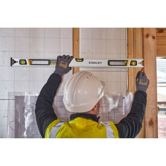 STANLEY FMHT43674-1 waterpas in gebruik op muur door werknemer met handschoenen en helm, bouwplaats.