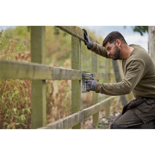 Agrafeuse STANLEY FMHT6-70868 utilisée sur clôture grillagée en extérieur, ouvrier avec gants, vue latérale.