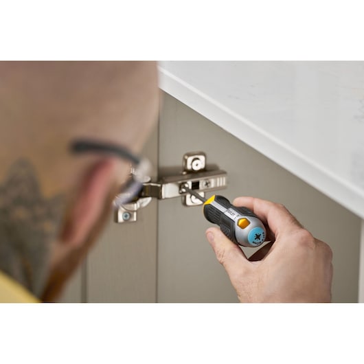 Une personne utilise un tournevis STANLEY pour serrer une charnière de meuble sous un plan de travail blanc.