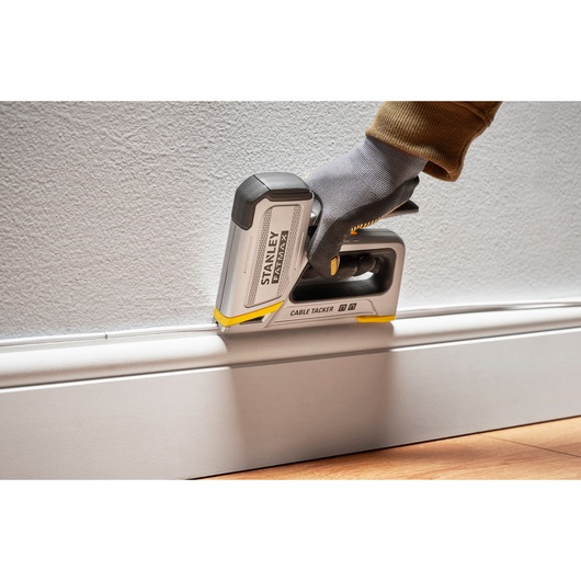 Stanley FMHT70100-0 agrafeuse de câbles utilisée le long d'une plinthe, tenue par une main gantée contre le mur.