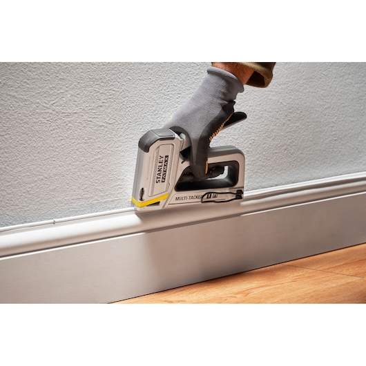 Photographie de l’agrafeuse multi-tacker STANLEY FMHT70250-0 utilisée, main gantée contre une plinthe.