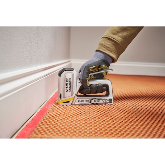 Agrafeuse multi-usage STANLEY FMHT70250-0 utilisée sur une moquette par une main gantée près d'une plinthe, photo en gros plan.
