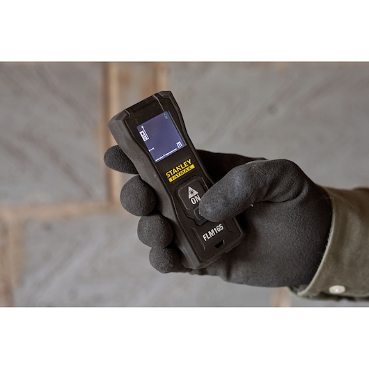 Photo du télémètre laser STANLEY FMHT77165-0 tenu dans une main gantée, écran et boutons visibles.