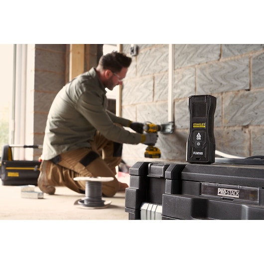 STANLEY FMHT77165-0 télémètre laser au premier plan sur une boîte PROSTACK, ouvrier en arrière-plan.