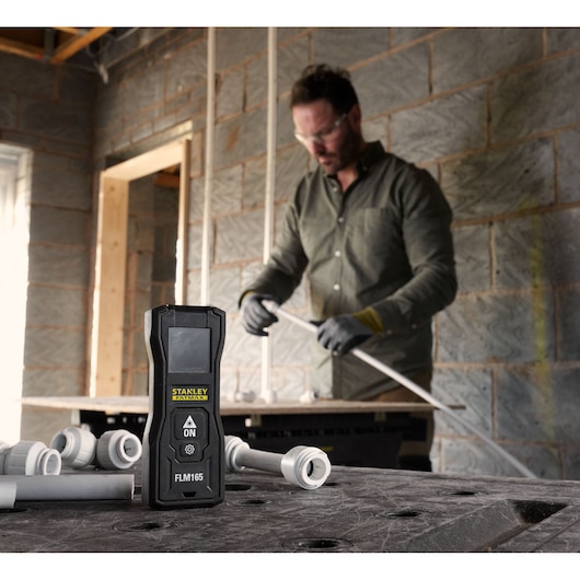 Photo du télémètre laser STANLEY FMHT77165-0 sur un établi de chantier avec tuyaux et raccords.