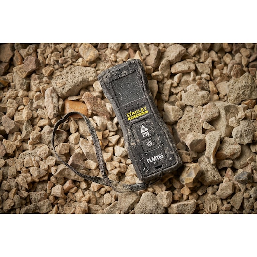 Télémètre laser STANLEY FMHT77165-0 avec dragonne sur sol rocheux, couvert de poussière et de gouttes d'eau.