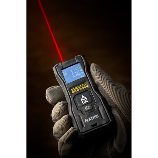STANLEY FMHT77165-0 télémètre laser dans une main gantée, écran affichant des mesures, faisceau laser visible.