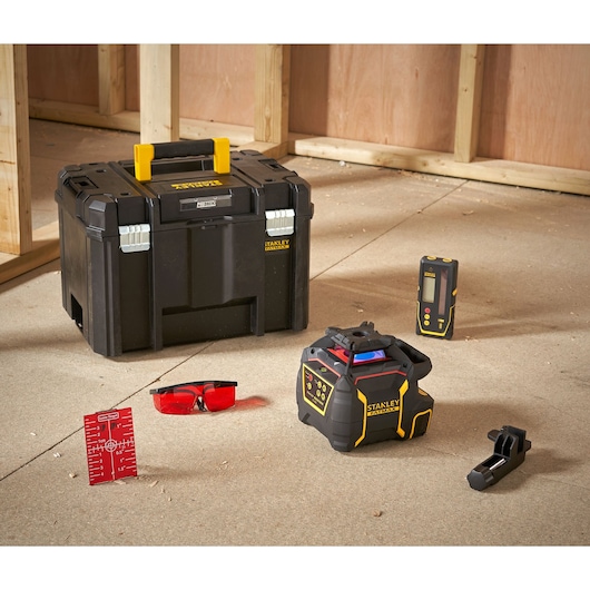 Ensemble d'outils STANLEY pour la photographie professionnelle et la mesure, comprenant une boîte à outils noire avec poignée jaune, un niveau laser, un détecteur laser, une pince, des lunettes de sécurité à verres rouges et une feuille cible rouge, disposés sur le sol d'un chantier avec une structure en bois en arrière-plan.
