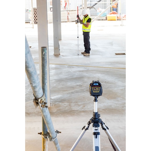 Niveau laser rotatif STANLEY FMHT77447-1 sur trépied sur chantier, ouvrier avec équipement de sécurité en arrière-plan.