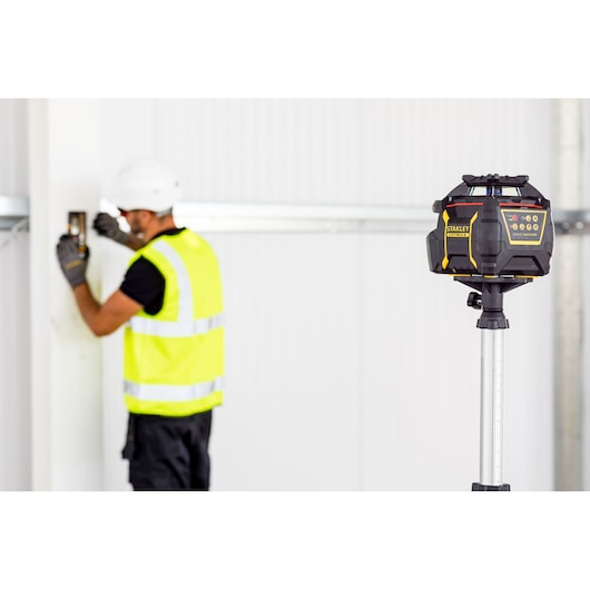 Niveau laser rotatif STANLEY FMHT77447-1 sur trépied, chantier, ouvrier en arrière-plan.